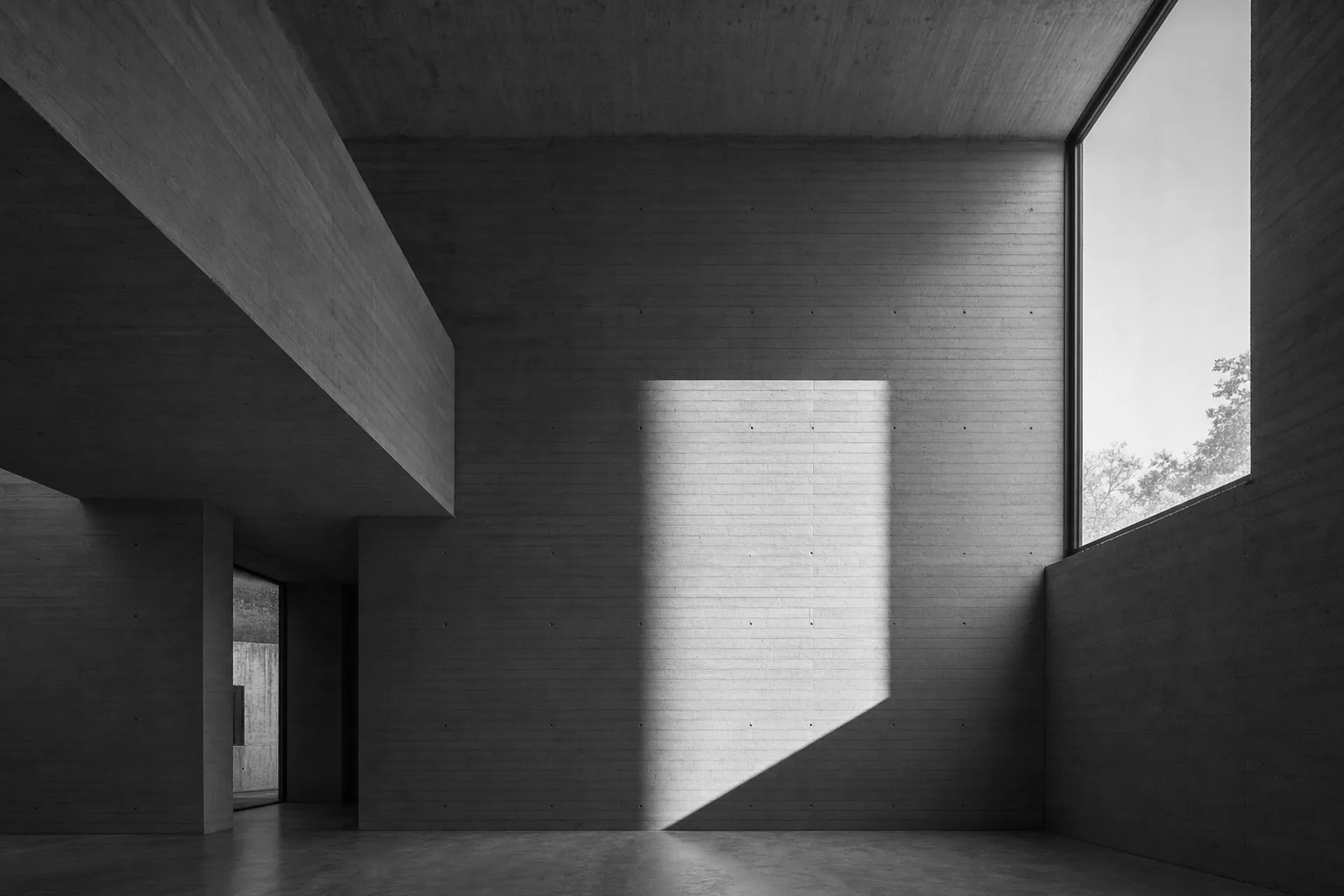 Intérieur de béton brut en double hauteur, une grande baie découpe un rectangle de lumière dure sur le mur banché.