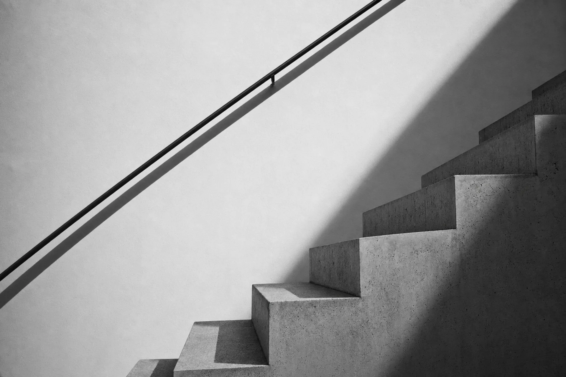 Détail d'un escalier de béton contre un mur blanc, ombre diagonale nette, main courante d'une seule ligne.