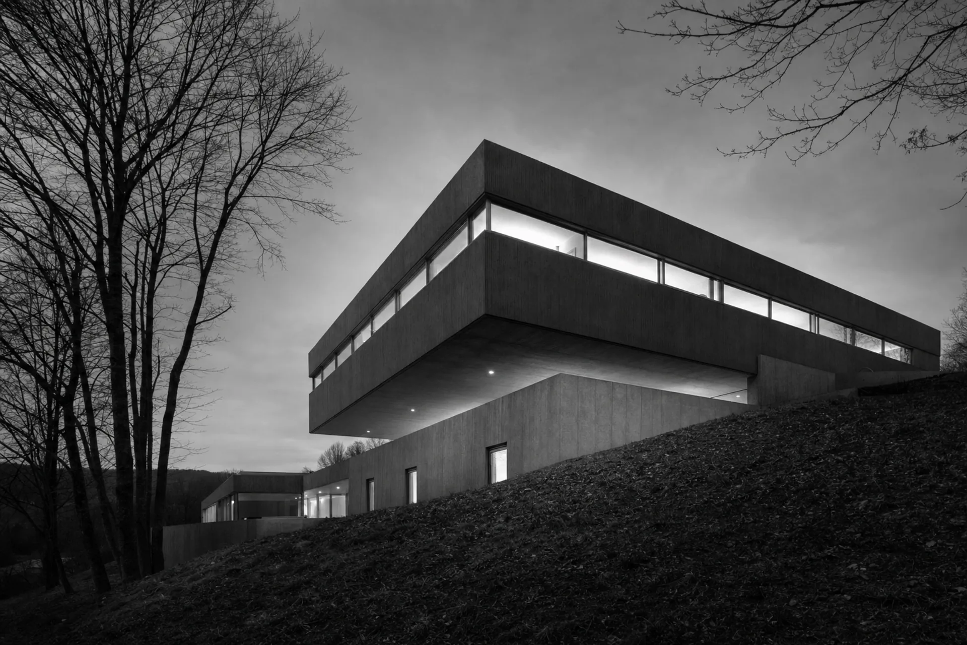 Maison moderniste en béton au crépuscule, volume en porte-à-faux au-dessus d'une pente.